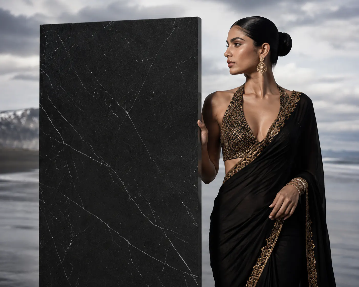A single black marble-look ORKAY slab stood on a black-sand shore, a woman in a black and gold sari beside it